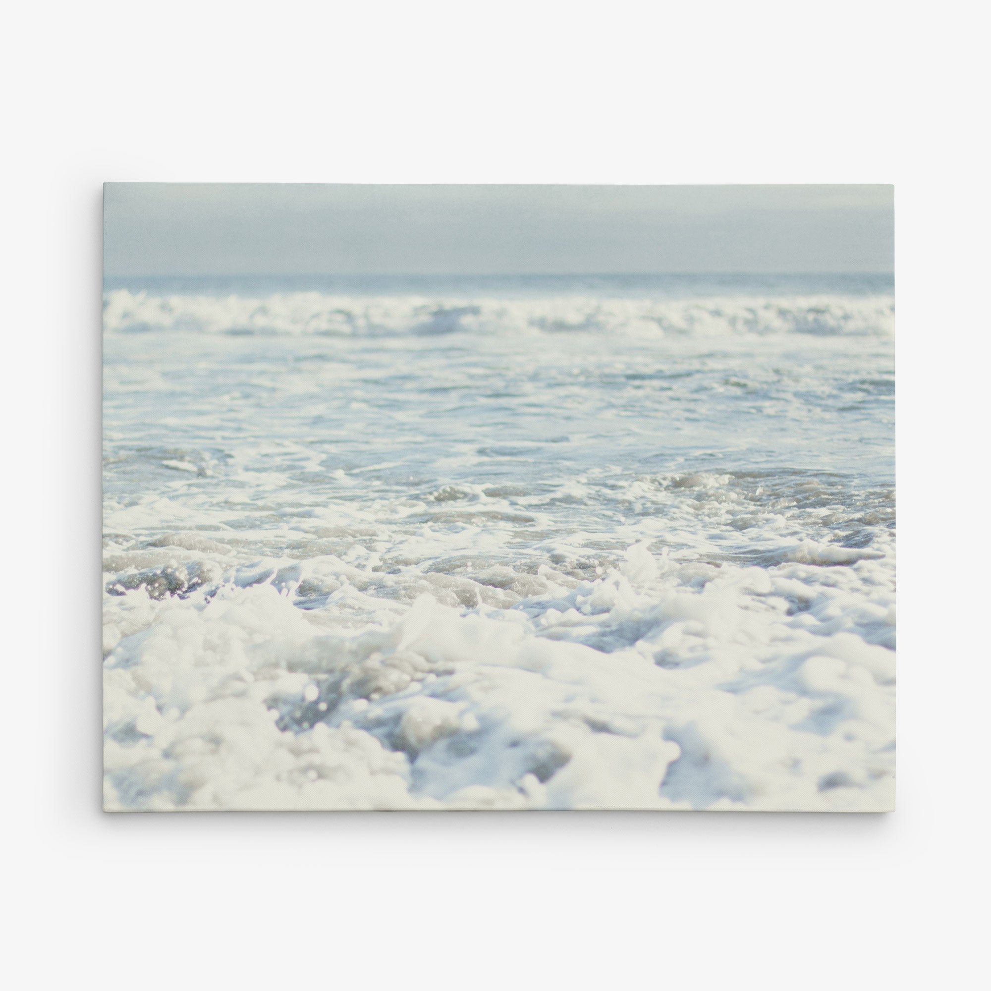 A serene ocean scene with gentle waves creating white foam as they approach the shore. The horizon is visible in the distance under a clear sky, blending with the calm sea. Ideal for coastal prints, Offley Green&#39;s 11x14 Coastal Canvas Print (Choose from 10+ Designs) is ready to hang and enhance any space with its tranquil beauty.