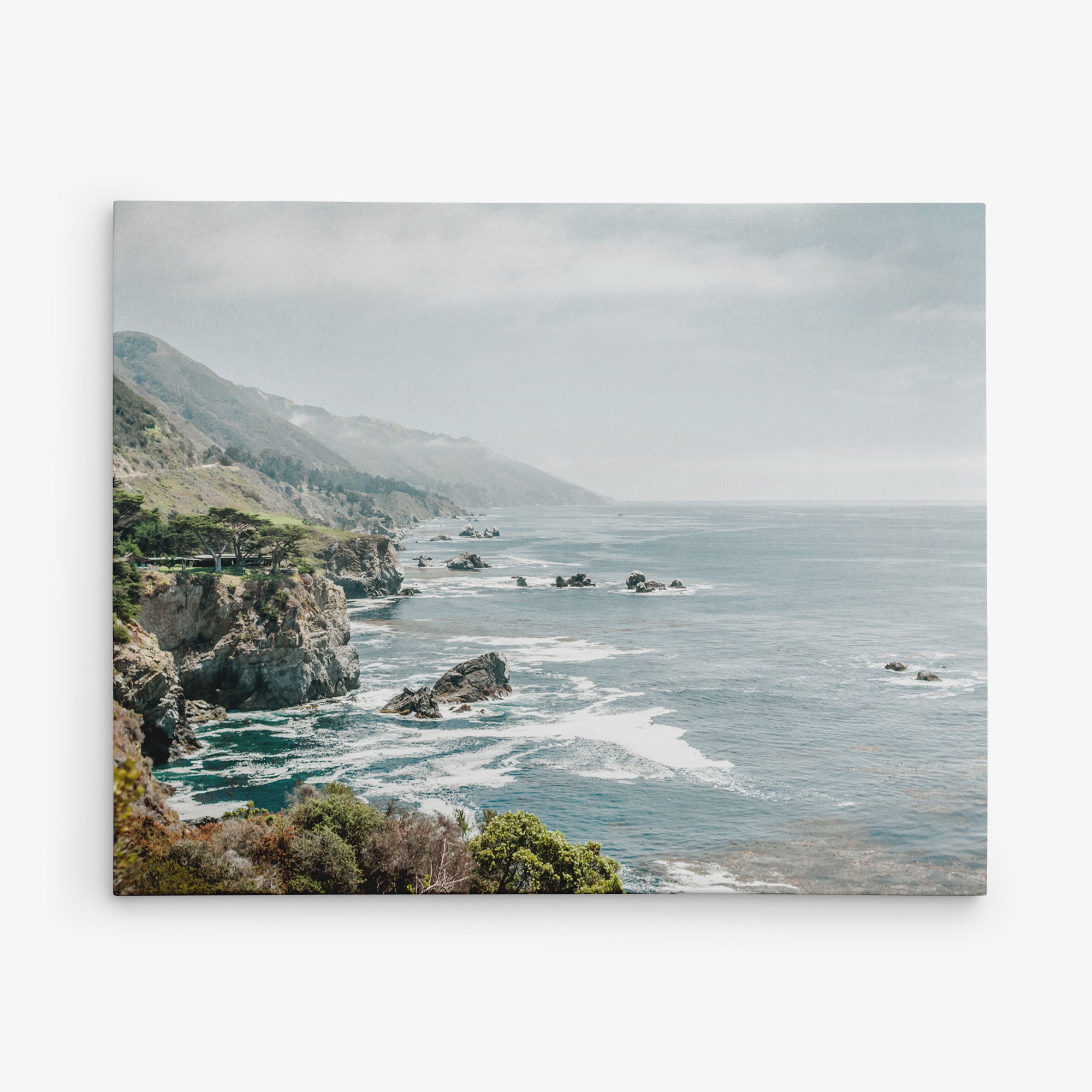 Experience the serene beauty of a rugged shoreline with cliffs and rocky outcrops extending into the ocean captured on the 11x14 Coastal Canvas Print from Offley Green. The calm water with gentle waves and a hint of mist over distant mountains and hills provides a picturesque scene, while lush vegetation adds greenery to the landscape. Choose from 10+ designs for the perfect coastal print to enhance your space.