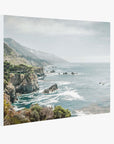 The Offley Green Big Sur Landscape Canvas Wall Art, 'Rocky Rocks,' showcasing a scenic coastal view with rocky cliffs and rugged terrain meeting the ocean. Waves crash against the shore, and misty mountains rise in the background under a partially cloudy sky. The foreground is filled with coastal vegetation, captured on premium artist-grade canvas.