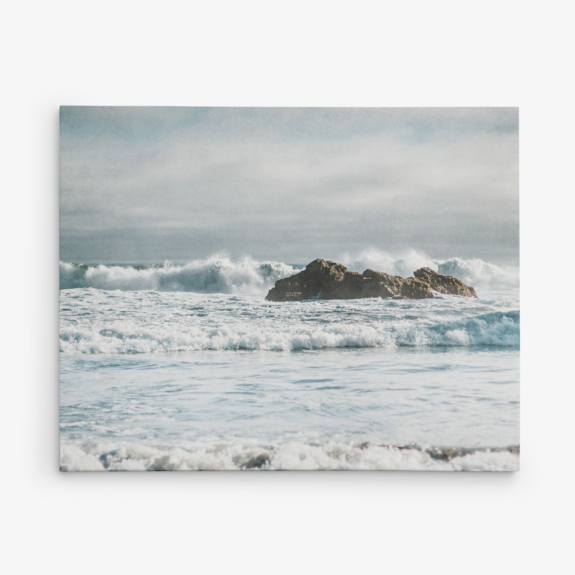 A rocky outcrop is surrounded by ocean waves, which appear rough and choppy. The horizon is somewhat obscured by a mist or haze, giving the scene a serene yet powerful ambiance. The sky above is mostly cloudy, adding to the dramatic mood of this seascape—a perfect subject for Offley Green&#39;s 8x10 Coastal Canvas Print (Choose from 10+ Designs) as wall art.