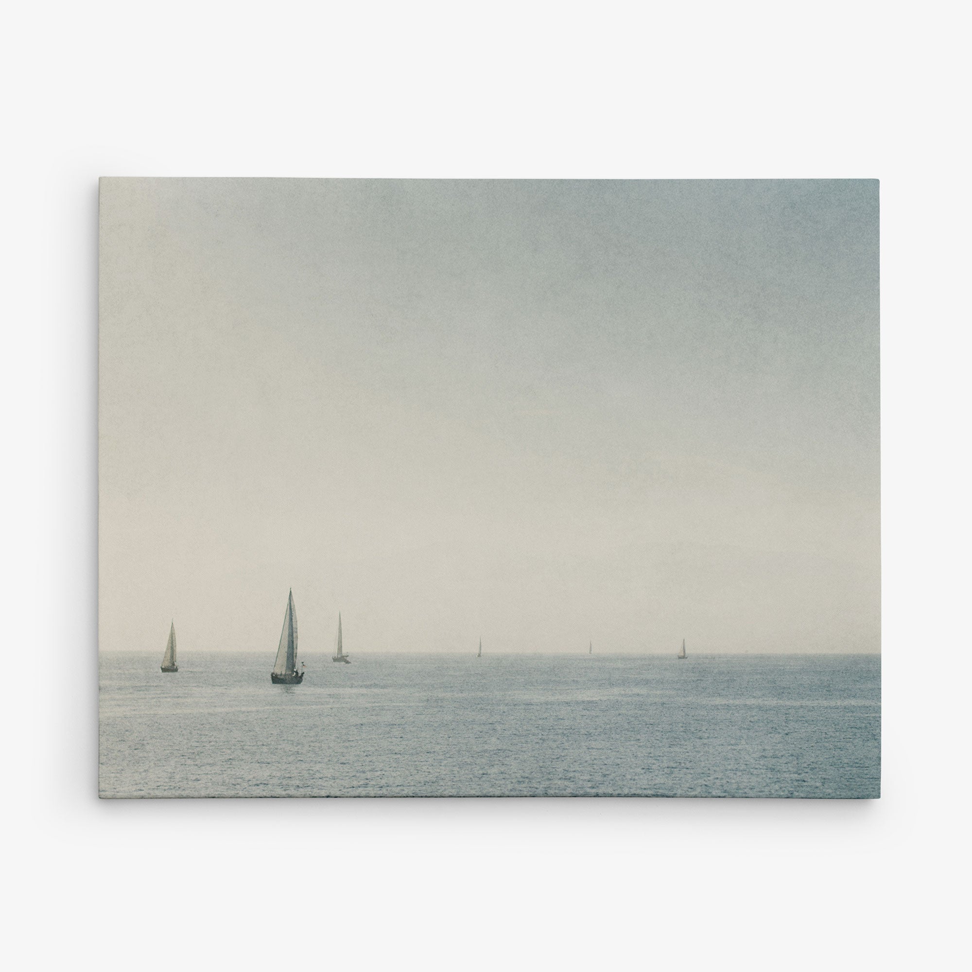 A serene seascape featuring several sailboats scattered across calm, open waters. The horizon blends softly with the sky, which is lightly overcast, casting a peaceful, muted light over the scene. This Offley Green &quot;11x14 Coastal Canvas Print&quot; (Choose from 10+ Designs) comes as a ready-to-hang canvas gallery wrap.