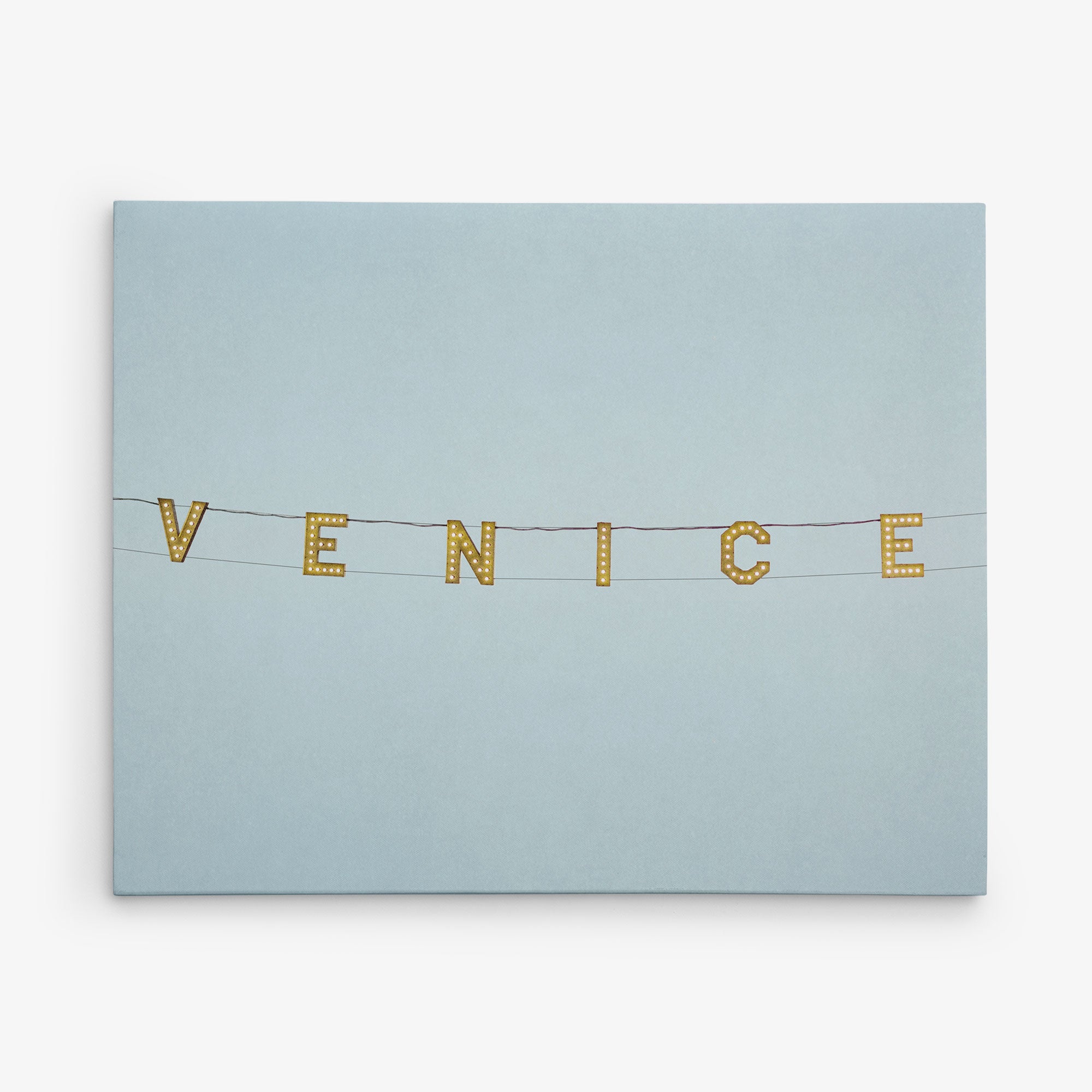 A clear sky with string lights spelling out &quot;VENICE&quot; hanging on black wires. The background is a gradient of light blue, creating a calm and serene atmosphere, reminiscent of Offley Green&#39;s 11x14 California Canvas Print (Choose from 10+ Designs) wall art or even a canvas gallery wrap.