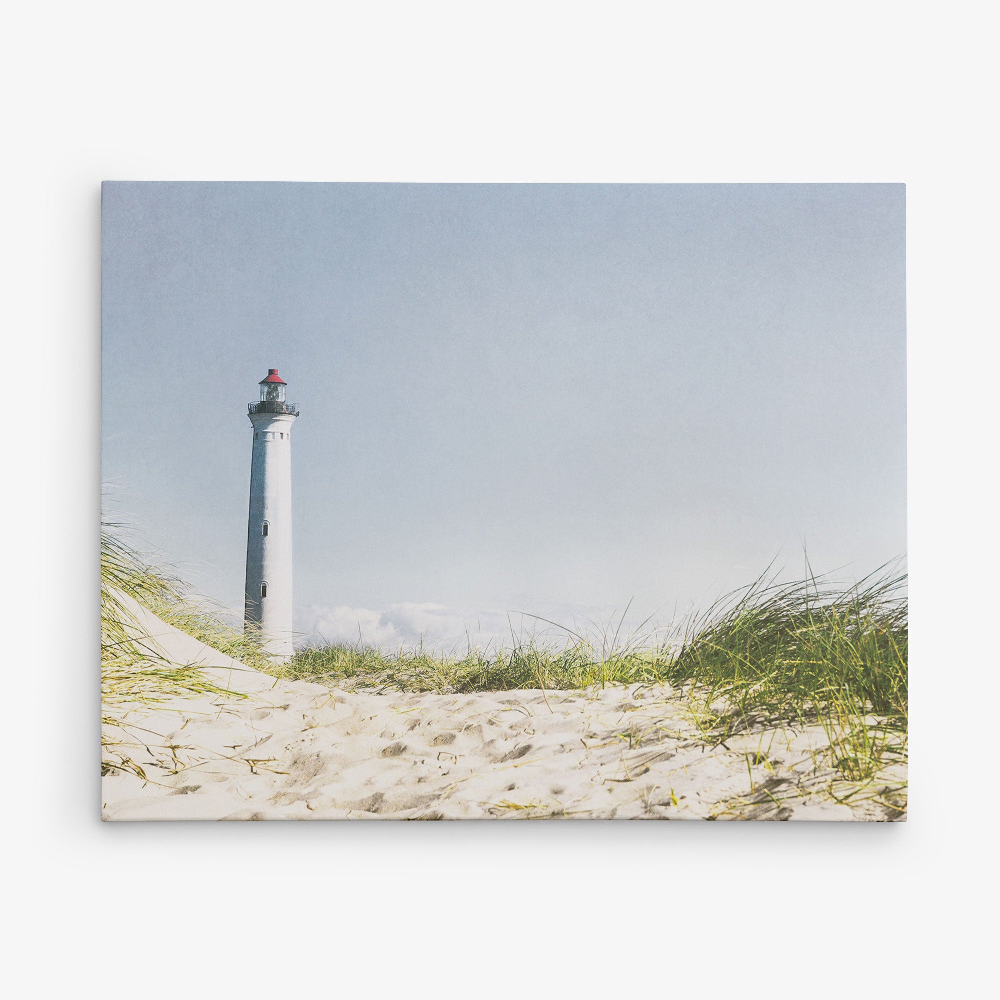 A serene 11x14 Coastal Canvas Print from Offley Green features a striking white lighthouse with a red top standing tall against a clear blue sky, surrounded by lush green grass and sandy dunes. This picturesque scene, one of the many designs available, evokes a calm ambiance and is ready to hang as a canvas gallery wrap in your home.