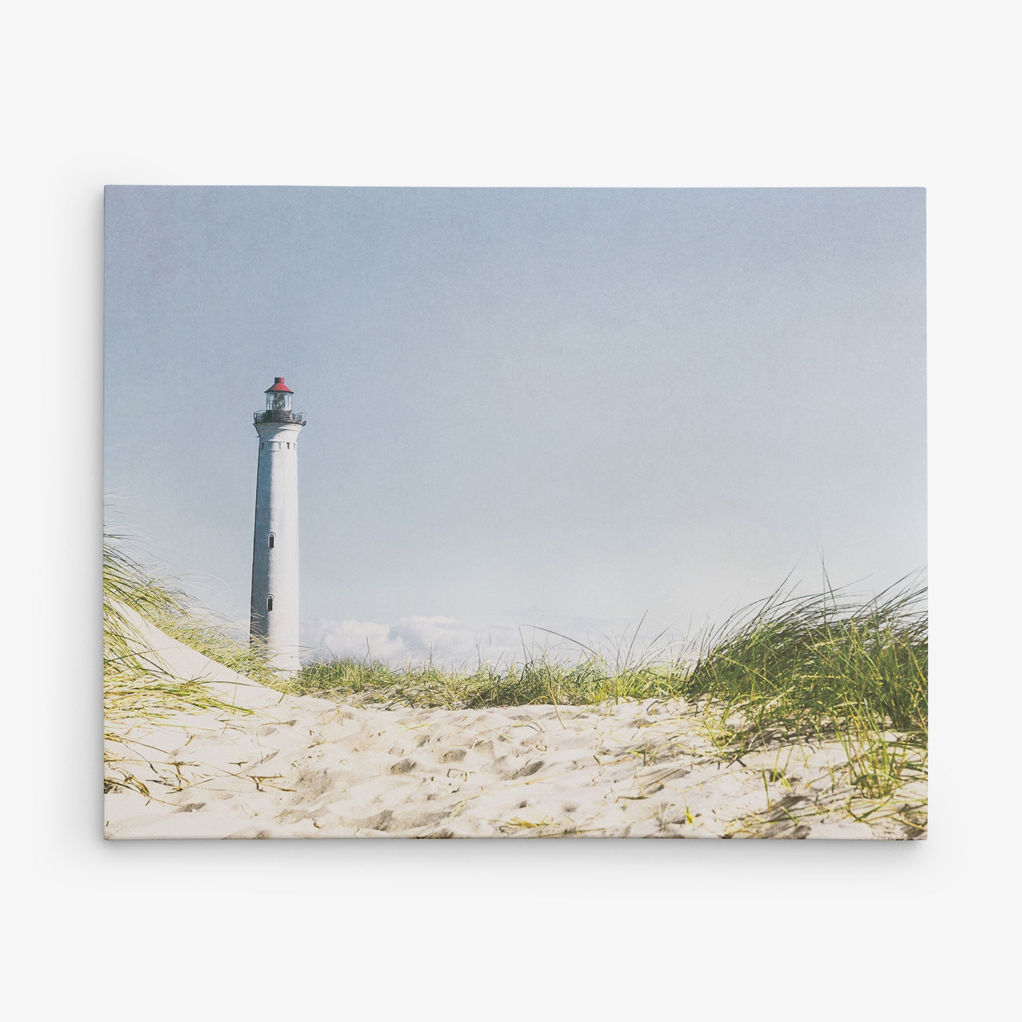 A tall, white lighthouse with a red top stands against a clear blue sky. The foreground features sand dunes covered with green grass. This bright and sunny scene evokes a peaceful coastal atmosphere, perfect for wall art or a canvas gallery wrap to bring seaside serenity to your space. Add this enchanting view from the Offley Green 8x10 Coastal Canvas Print collection and choose from more than 10 stunning designs.