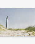 A serene 11x14 Coastal Canvas Print from Offley Green features a striking white lighthouse with a red top standing tall against a clear blue sky, surrounded by lush green grass and sandy dunes. This picturesque scene, one of the many designs available, evokes a calm ambiance and is ready to hang as a canvas gallery wrap in your home.