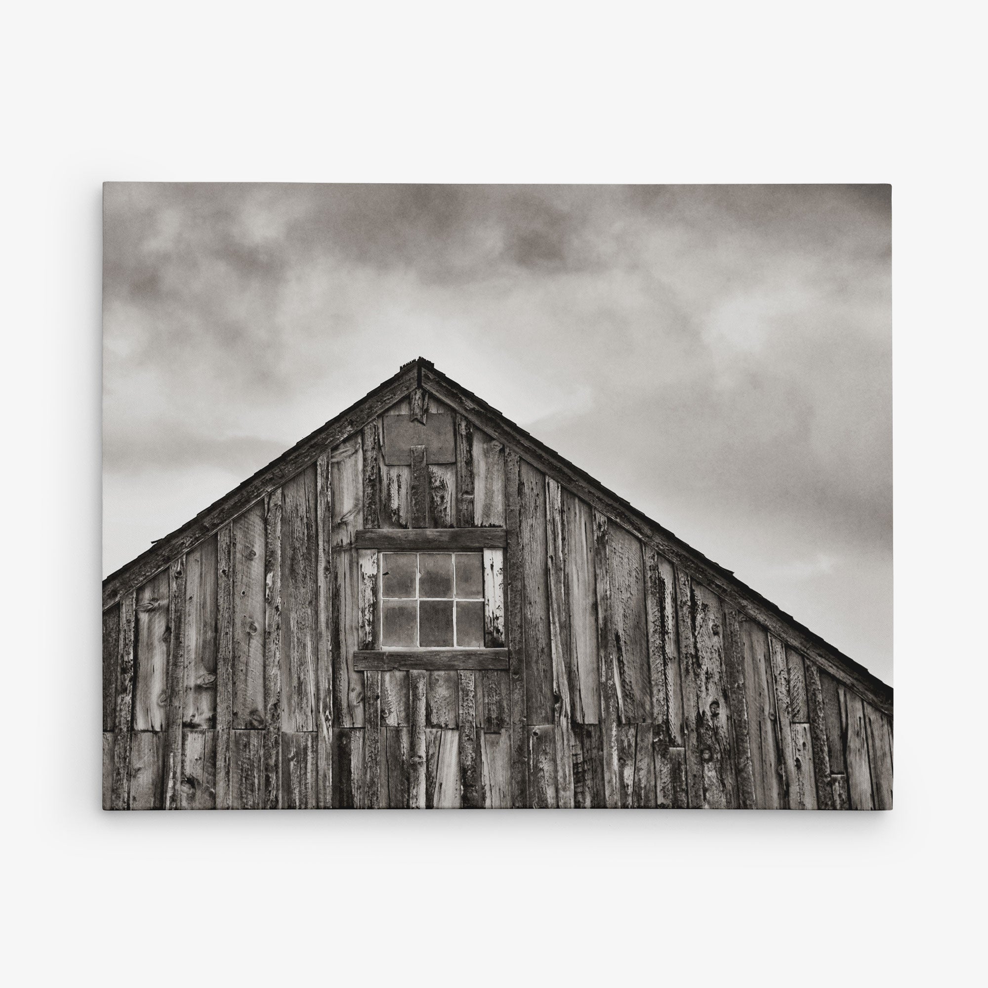 Black and white image of an old wooden barn&#39;s upper section against a cloudy sky. The barn has weathered wood planks and a small, rectangular window at the center of the triangular peak. This piece of rustic wall art evokes nostalgia, making it perfect for a 16x20 Rustic Canvas Print (Choose from 10+ Designs) by Offley Green.