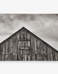 A black-and-white image of the top half of an old wooden barn. The barn is weathered, with vertical wooden planks and a window centered beneath the triangular roof. The sky is cloudy, giving a moody atmosphere to this Offley Green 30x40 Rustic Canvas Print (Choose from 10+ Designs).