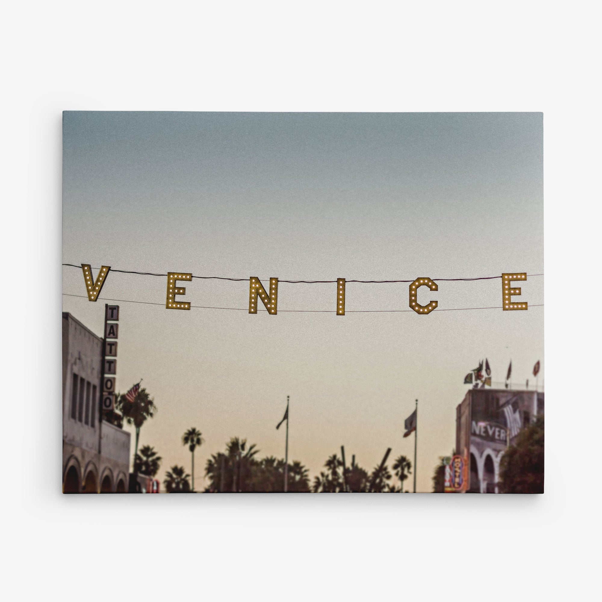 A photo of the Venice sign in Venice Beach, California. The illuminated letters hang across the street at sunset, capturing the essence of this iconic locale. Palm trees and buildings, including a tattoo shop and a &quot;NEVER&quot; sign, add to the charm—perfect for Offley Green&#39;s 16x20 California Canvas Print (Choose from 10+ Designs).