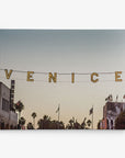 The image displays a sunset street view with the word "VENICE" spelled out in large, illuminated letters hanging above. Palm trees, buildings, and a tattoo shop sign are visible in the background, making this scene ideal for California-themed prints. This artwork is available as a 30x40 California Canvas Print from Offley Green, which offers over 10 different designs to choose from.