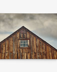 A photograph of the upper portion of a rustic wooden barn with a single window centered near the peak of its gabled roof. The weathered wood panels display various shades of brown and gray, perfect for an Offley Green 16x20 Rustic Canvas Print (Choose from 10+ Designs). The sky above is overcast with a mix of dark and light clouds.