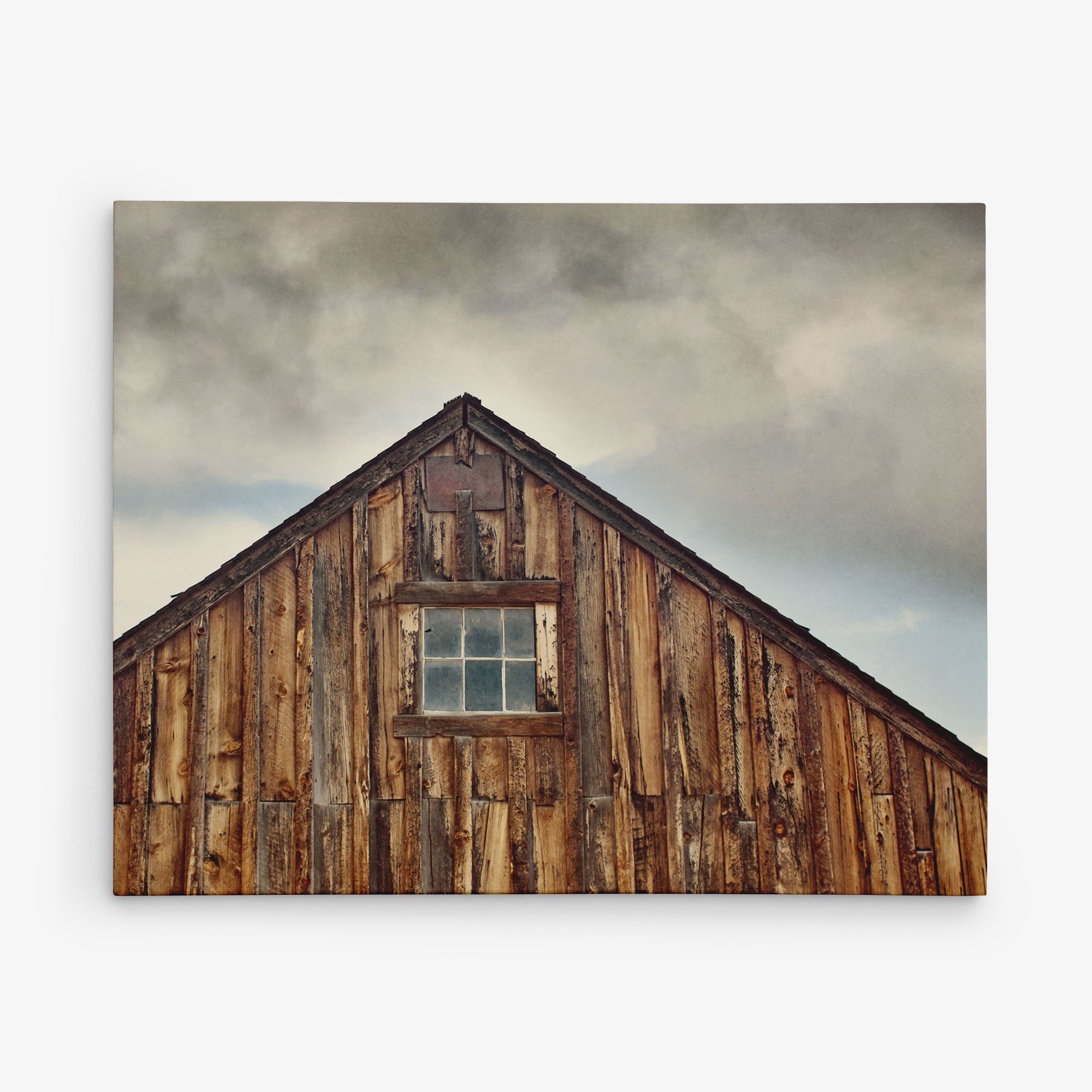 A rustic, weathered wooden barn with a steeply pitched roof stands against a cloudy sky. A single square window is centered near the roof&#39;s peak, showcasing the barn&#39;s aged, textured wood planks—perfect for transforming into Offley Green&#39;s 24x30 Rustic Canvas Print (Choose from 10+ Designs) on premium artist-grade canvas.