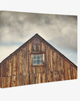 An image of a weathered, wooden barn with a single rectangular window near the peak of the roof. The sky above is overcast with thick clouds. The barn's wooden planks display varying tones of brown and visible aging—perfect as an 11x14 Rustic Canvas Print (Choose from 10+ Designs) from Offley Green.