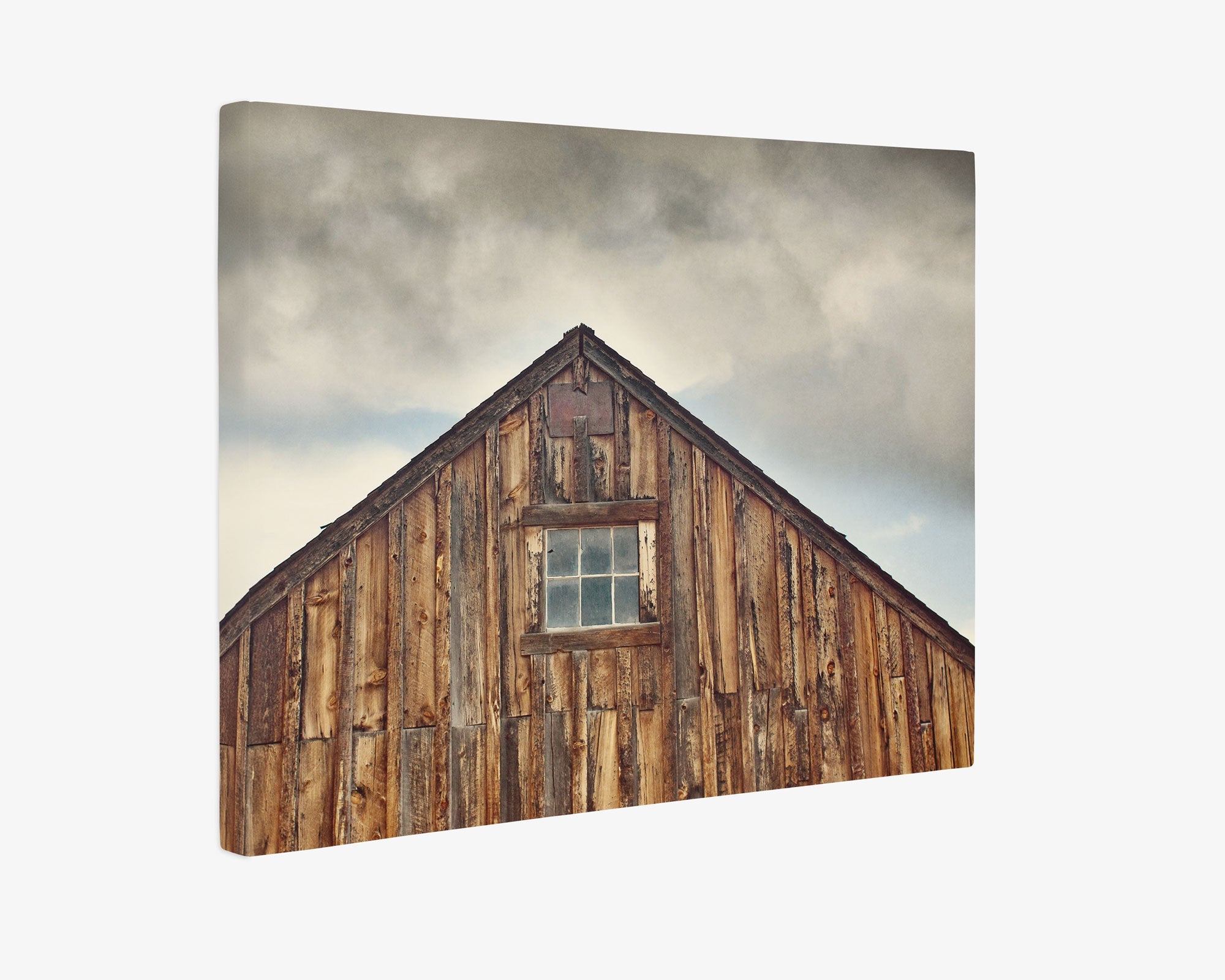 A 16x20 Rustic Canvas Print (Choose from 10+ Designs) by Offley Green, featuring a wooden barn with vertical planks and a single square window at the center of its triangular gable, evokes the essence of rustic wall art. The sky above is overcast with dark, moody clouds, enhancing its weathered atmosphere.