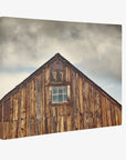 A close-up view of the upper section of a rustic wooden barn with a single square window in the center. The barn's wood is weathered and aged, and the sky above is overcast, giving a moody atmosphere, perfect for capturing as Offley Green 24x30 Rustic Canvas Print (Choose from 10+ Designs).