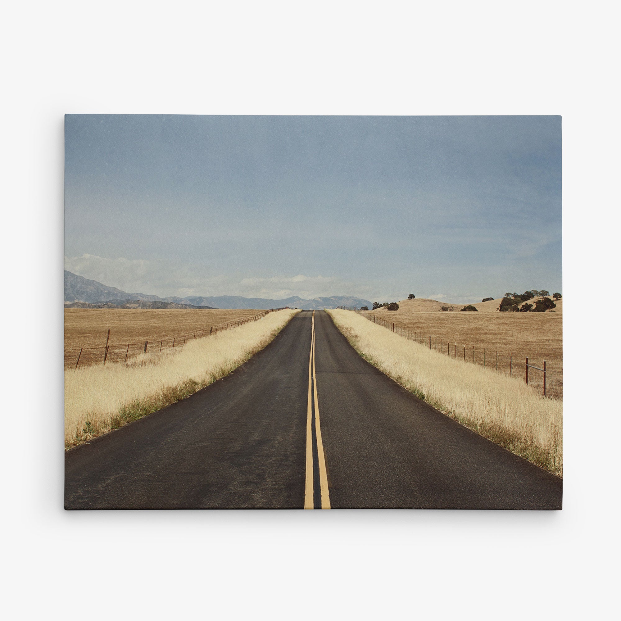 A straight, empty road stretches into the horizon with a double yellow line running down the center. The road is bordered by dry, golden grasses and fenced fields, under a vast, clear blue sky. Distant mountains are visible at the far end of the road. Perfect as 11x14 Rustic Canvas Print (Choose from 10+ Designs) by Offley Green for wall art or canvas gallery wraps.