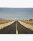 A straight, empty road stretches into the horizon with a double yellow line running down the center. The road is bordered by dry, golden grasses and fenced fields, under a vast, clear blue sky. Distant mountains are visible at the far end of the road. Perfect as 11x14 Rustic Canvas Print (Choose from 10+ Designs) by Offley Green for wall art or canvas gallery wraps.