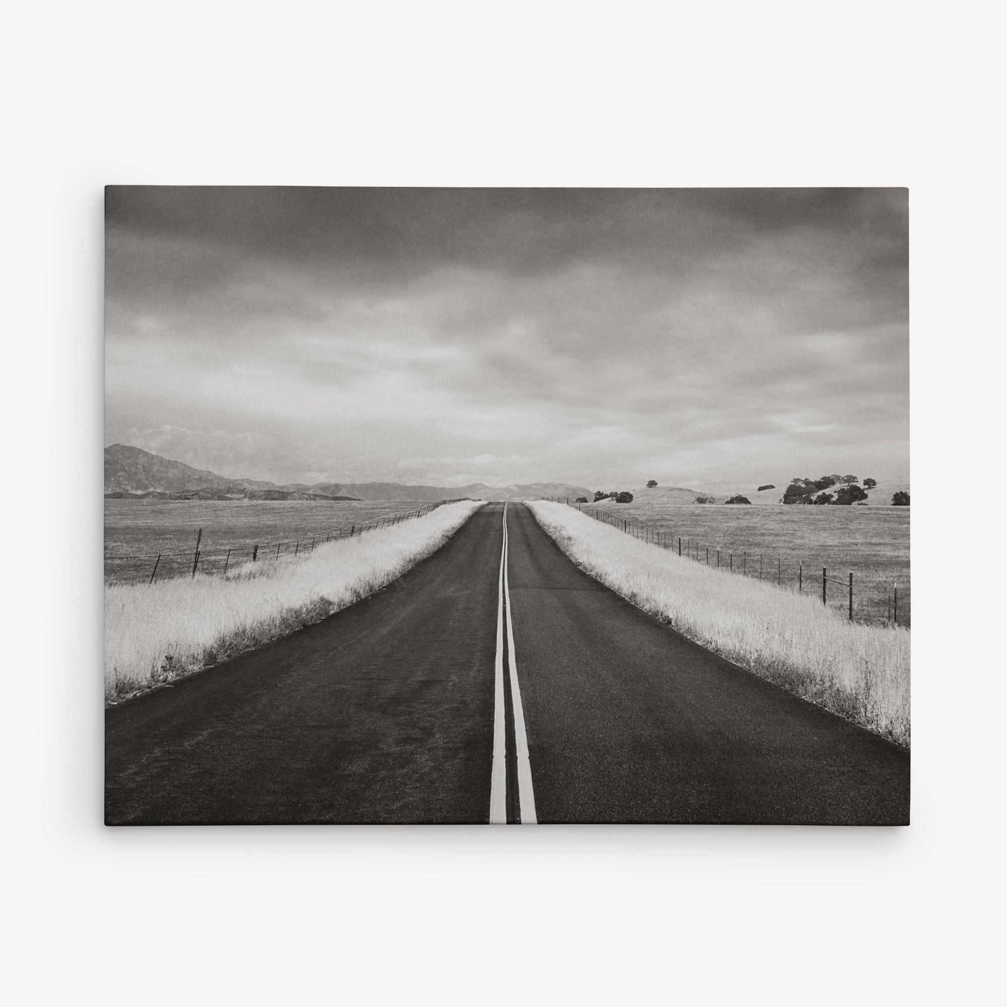 A black-and-white photograph of a straight road stretching into the distance, flanked by grassy fields and fences. The sky above is overcast, creating a moody atmosphere. In the far distance, mountains are faintly visible, making Offley Green&#39;s 11x14 Rustic Canvas Print (Choose from 10+ Designs) perfect wall art for those who love rustic prints.