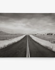 A black-and-white photograph of a straight road stretching into the distance, flanked by grassy fields and fences. The sky above is overcast, creating a moody atmosphere. In the far distance, mountains are faintly visible, making Offley Green's 11x14 Rustic Canvas Print (Choose from 10+ Designs) perfect wall art for those who love rustic prints.