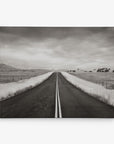 A black and white photo depicting a long, empty road that stretches into the horizon. The road is bordered by fields of grass and fenced farmland, with a cloudy sky overhead. This Offley Green 16x20 Rustic Canvas Print (Choose from 10+ Designs) symmetrical composition emphasizes the road's solitary and endless nature.