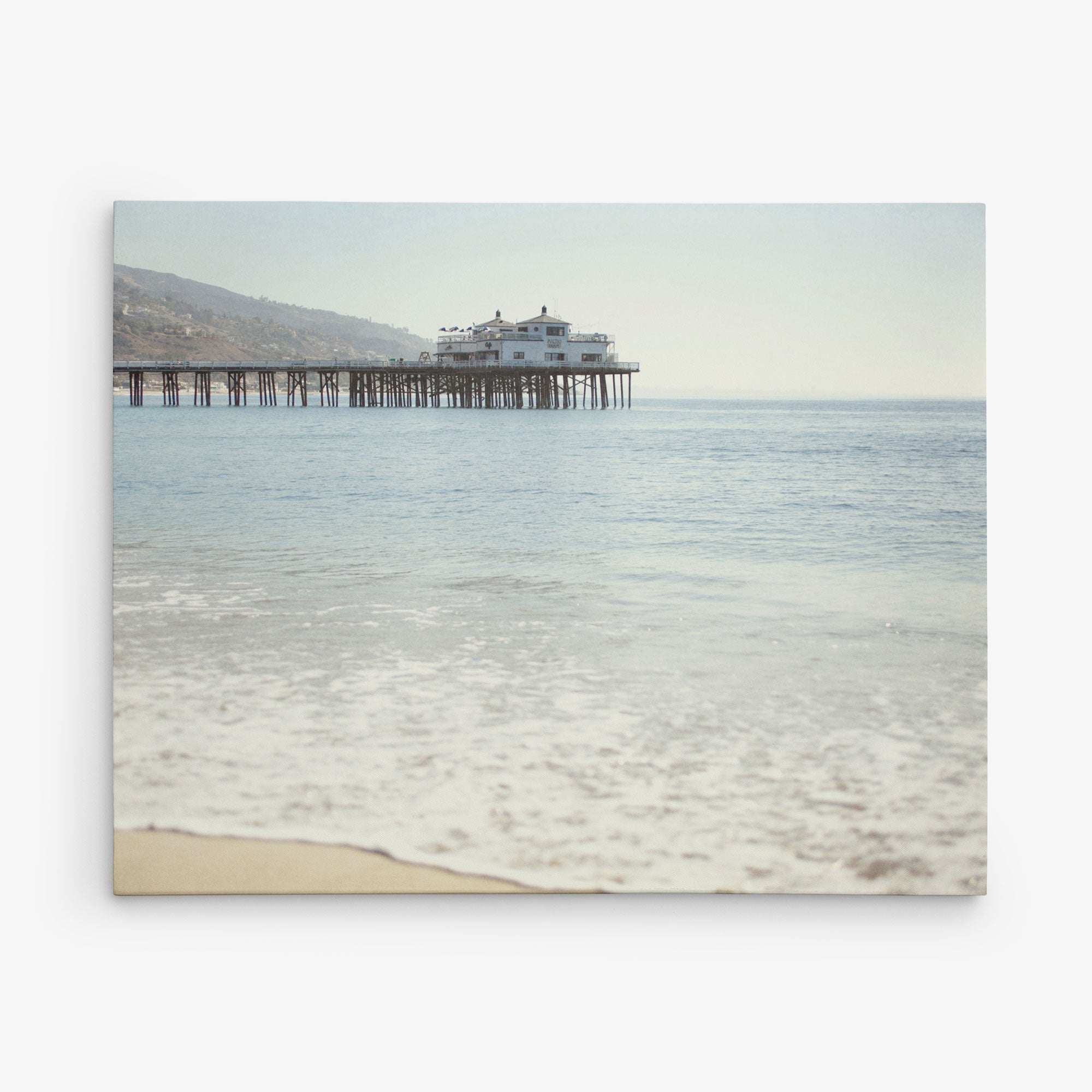 A tranquil beach scene featuring a long wooden pier extending into calm ocean waters graces this 8x10 California Canvas Print from Offley Green. The clear sky and faintly visible distant hills add to the serenity, as gentle waves wash onto the sandy shore. This stunning image, perfect for any of the 10+ designs available, would make beautiful wall art in any space.