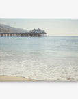 A serene beach scene featuring a calm ocean and a large pier extending into the water, with a building on the end of the pier. Gentle waves lap the sandy shore under a clear, blue sky. Hills are faintly visible in the background, making it perfect for an Offley Green 11x14 California Canvas Print (Choose from 10+ Designs) as stunning wall art.