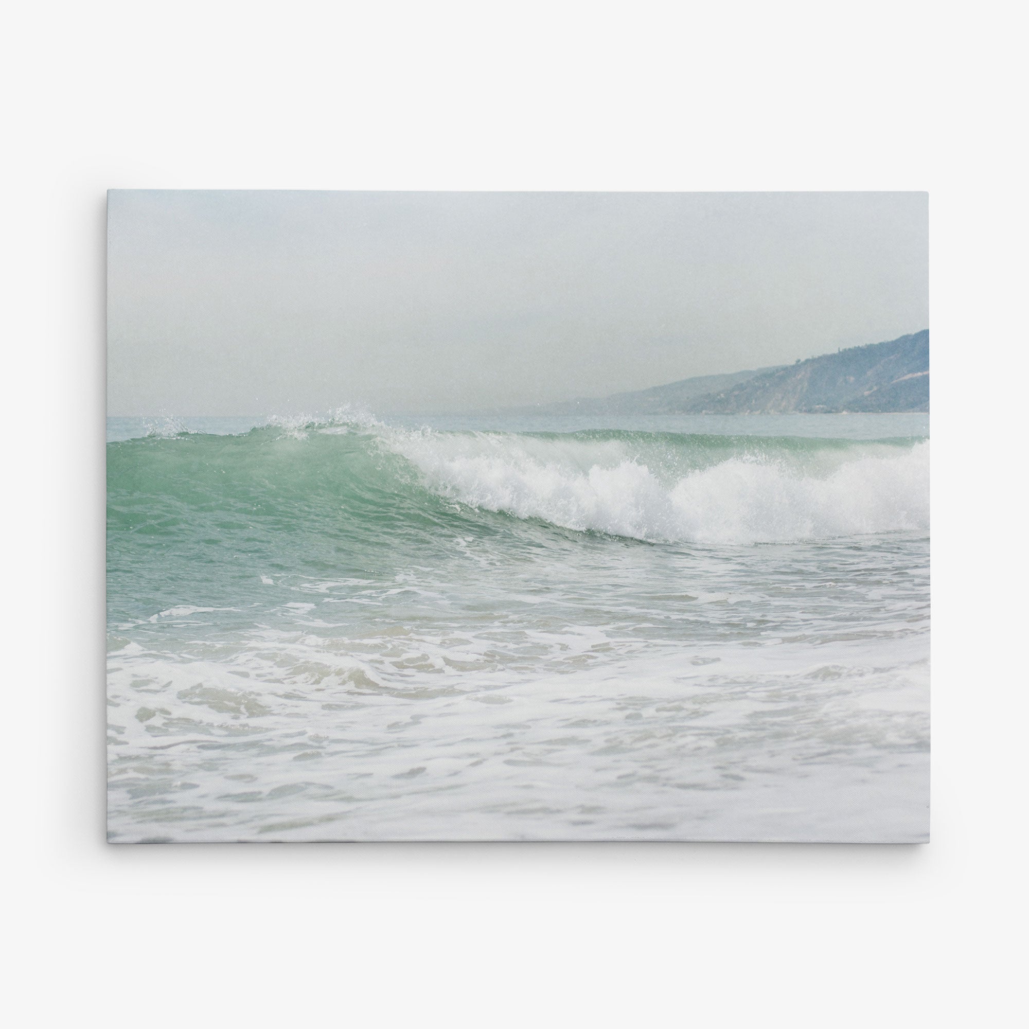 A serene coastal scene capturing a gentle wave rolling towards a sandy shore under an overcast sky. In the background, a misty hillside meets the horizon, blending seamlessly with the calm sea. Introducing the 11x14 Coastal Canvas Print from Offley Green—part of a collection featuring over 10 beautiful designs. These canvas gallery wraps are ready to hang and transform any space.