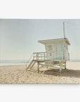 A solitary lifeguard tower stands on a sandy beach under a clear blue sky, perfect for Offley Green's 11x14 California Canvas Print (Choose from 10+ Designs). The tower has a small structure with windows, a door, and a set of stairs leading up to it. The ocean is visible in the background, and a safety sign is posted on the tower—ideal for wall art or canvas gallery wrap.