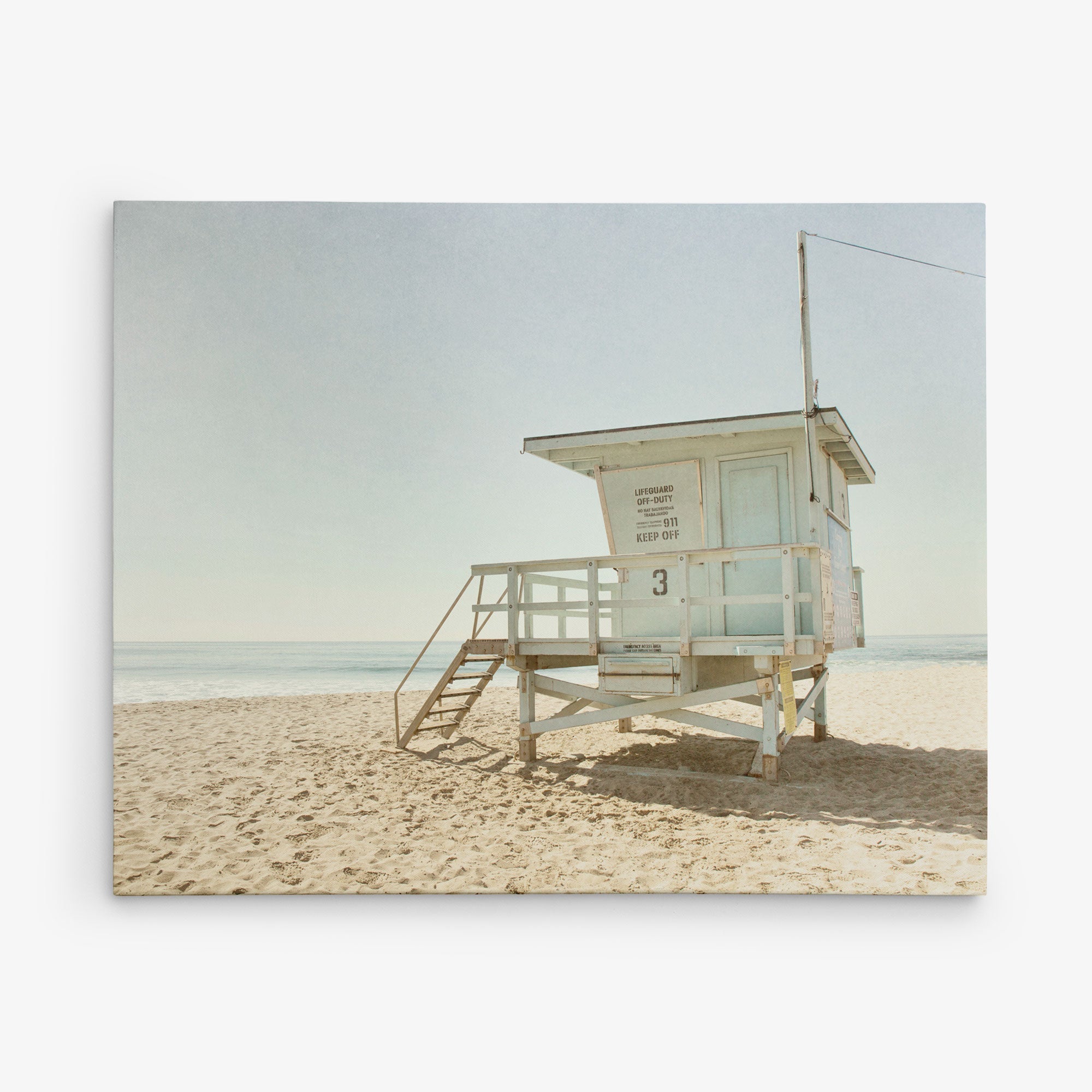 A solitary lifeguard tower stands on a sandy beach under a clear sky. The structure, painted in a weathered white, overlooks the calm ocean. The steps leading up to the platform are empty, and there are no people around, creating a tranquil scene reminiscent of Offley Green&#39;s 16x20 California Canvas Print (Choose from 10+ Designs).