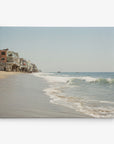 A serene beach scene with gentle waves lapping the sandy shore. Several beach houses line the left side of the image, elevated on stilts. The sky is clear, creating a tranquil atmosphere, perfect for wall art or an 11x14 California Canvas Print (Choose from 10+ Designs) by Offley Green to bring coastal calm to your home.