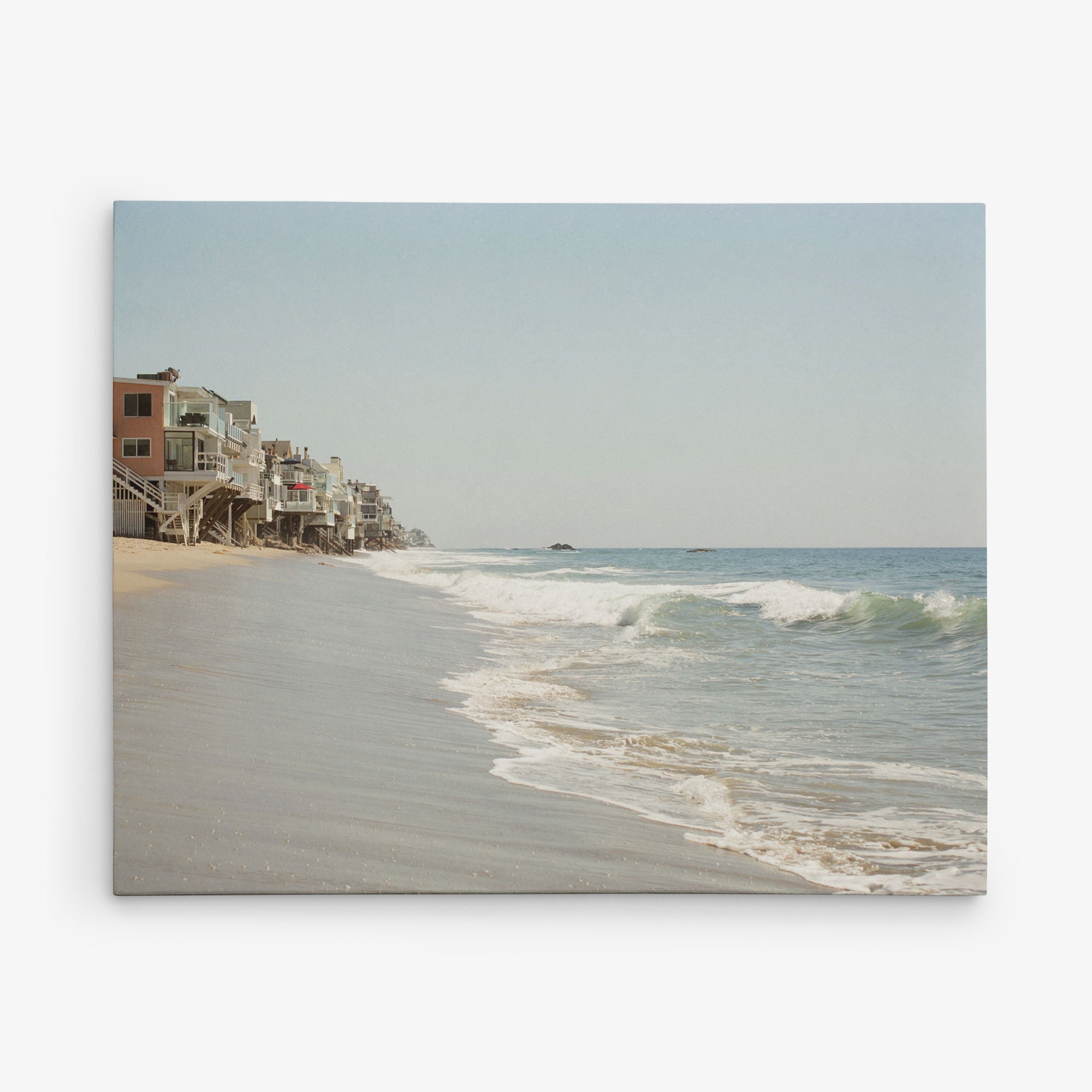 A tranquil beach scene with gentle waves lapping against the shore under a clear sky. Beachfront houses stand closely together on stilts along the left side. The sandy shoreline extends far into the distance, gradually fading toward the horizon—an ideal subject for the 8x10 California Canvas Print by Offley Green (Choose from 10+ Designs).