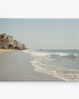 A tranquil beach scene with gentle waves lapping against the shore under a clear sky. Beachfront houses stand closely together on stilts along the left side. The sandy shoreline extends far into the distance, gradually fading toward the horizon—an ideal subject for the 8x10 California Canvas Print by Offley Green (Choose from 10+ Designs).