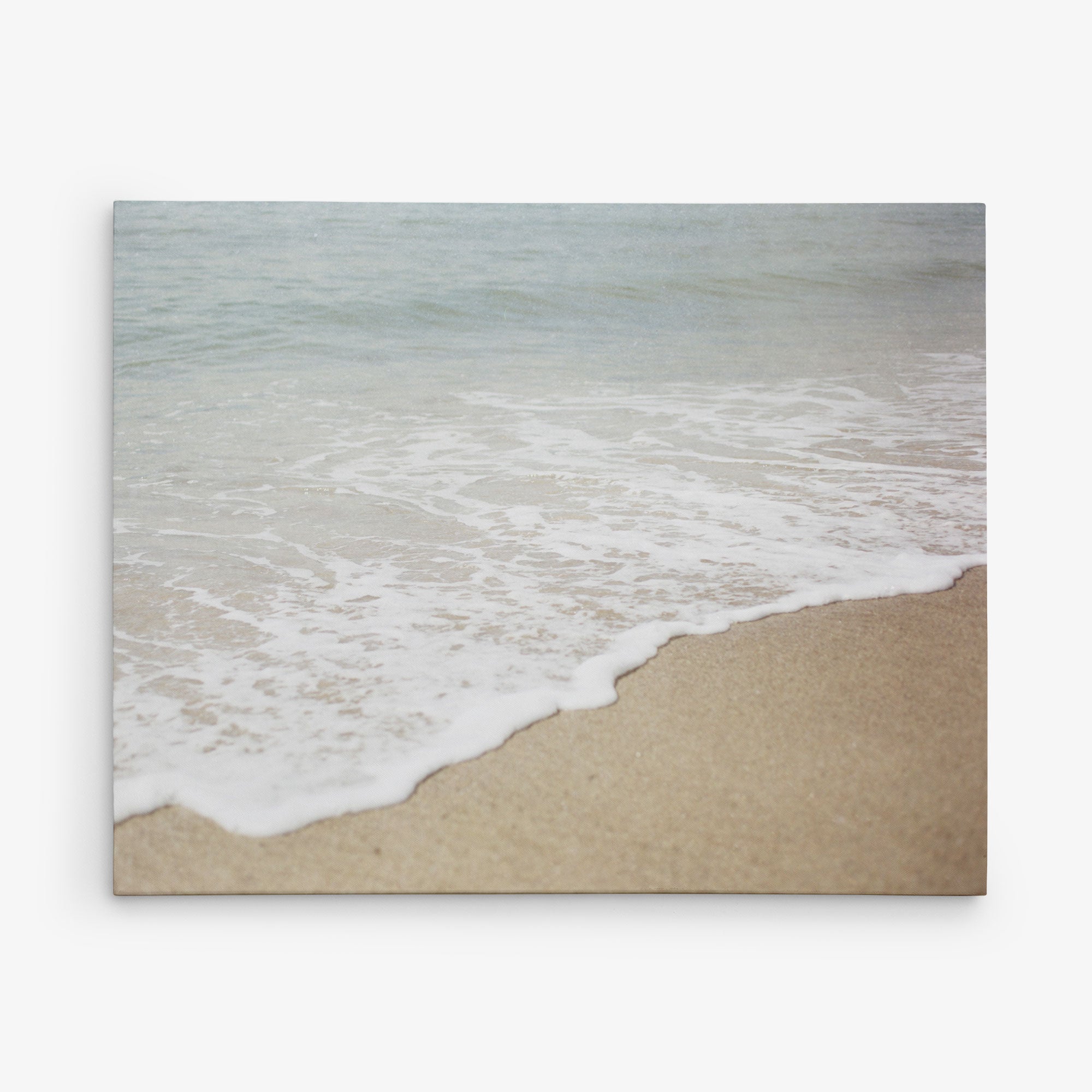 A tranquil beach scene showcasing gentle ocean waves softly rolling onto a sandy shore. The pale blue water seamlessly merges with the beige sand, fostering a serene and peaceful ambiance. Ideal as coastal prints or on an Offley Green 24x30 Coastal Canvas Print (Choose from 10+ Designs) to bring tranquility to any space.