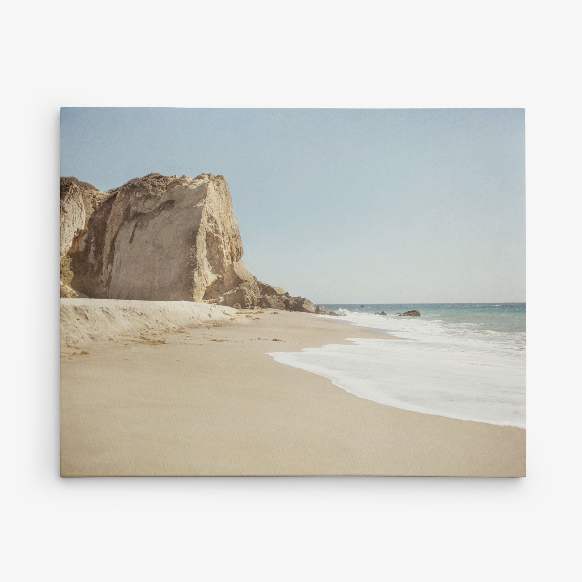 A tranquil beach scene with gentle waves washing onto a sandy shore. To the left, a steep, rugged cliff rises against a clear blue sky. The horizon meets the sea in the distance, creating a serene and picturesque view perfect for Offley Green’s 30x40 Coastal Canvas Print (Choose from 10+ Designs).