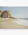 A tranquil beach scene with gentle waves washing onto a sandy shore. To the left, a steep, rugged cliff rises against a clear blue sky. The horizon meets the sea in the distance, creating a serene and picturesque view perfect for Offley Green’s 30x40 Coastal Canvas Print (Choose from 10+ Designs).