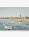 A scenic beach view shows waves crashing onto the shore, where numerous people are enjoying the sun and sand. In the background, a cityscape with tall buildings stretches along the coastline, with mountains visible in the distance under a clear sky—a perfect scene for an Offley Green 30x40 California Canvas Print (Choose from 10+ Designs).