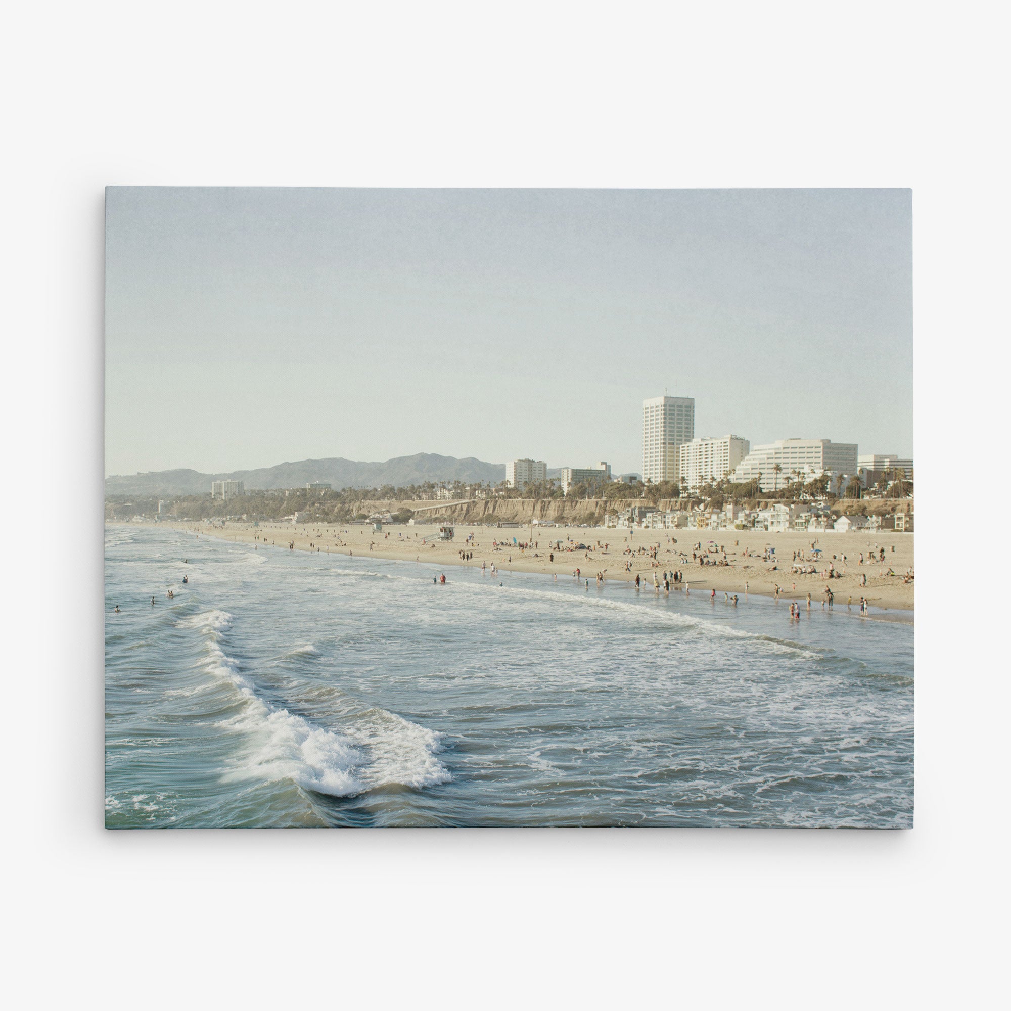 A wide view of a sandy beach with gentle waves crashing on the shore. The beach is dotted with people enjoying the sun and sea. In the background, buildings and palm trees stand tall, with mountains faintly visible in the distance. This scene would make stunning Offley Green 11x14 California Canvas Print (Choose from 10+ Designs).