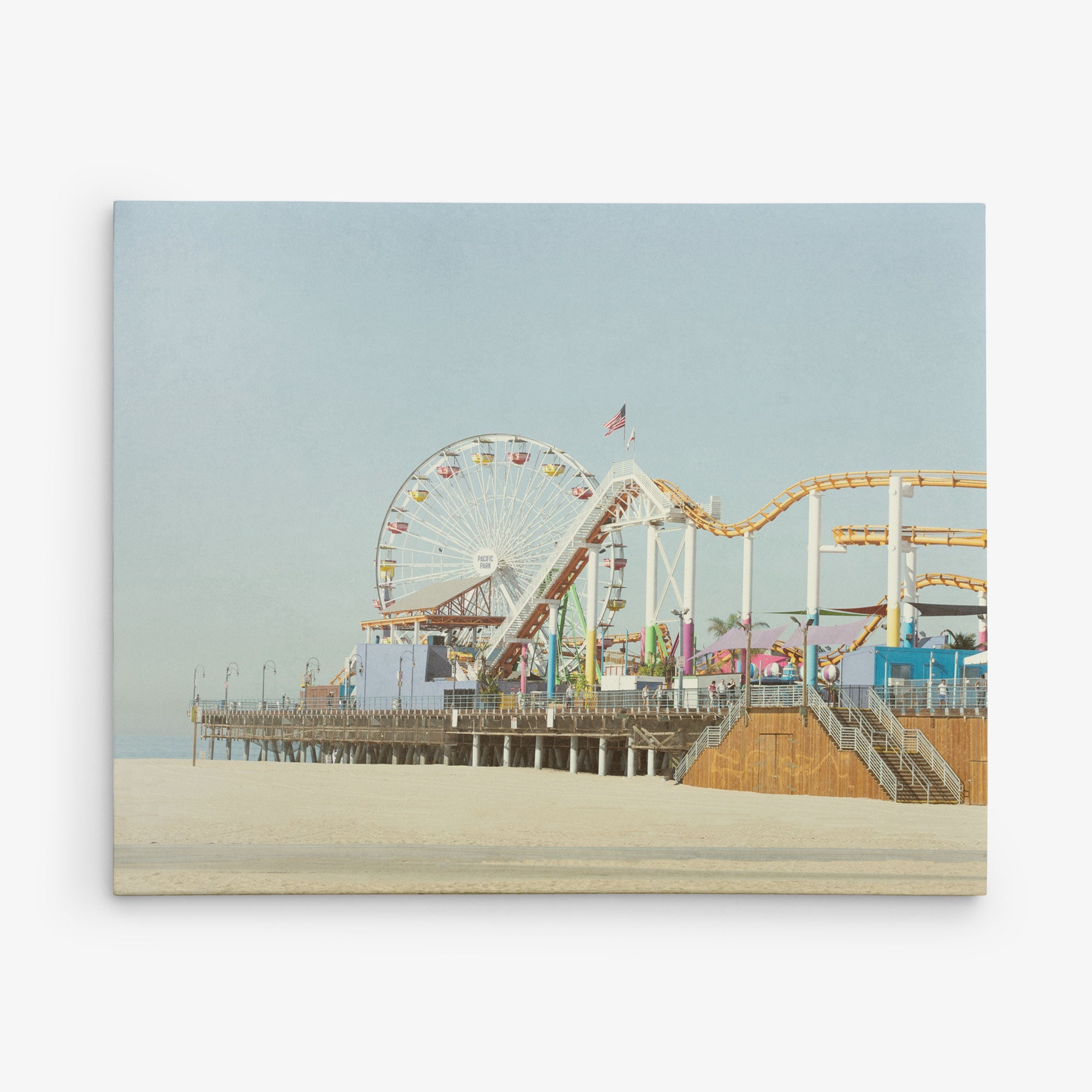 An image of a sunny beach boardwalk featuring a colorful Ferris wheel, roller coaster, and other amusement park rides on a pier. The background displays a clear, pale blue sky and stretches of sandy beach in the foreground—perfect for Offley Green&#39;s 8x10 California Canvas Print (Choose from 10+ Designs), capturing the essence of California prints.