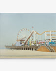 An image of a sunny beach boardwalk featuring a colorful Ferris wheel, roller coaster, and other amusement park rides on a pier. The background displays a clear, pale blue sky and stretches of sandy beach in the foreground—perfect for Offley Green's 8x10 California Canvas Print (Choose from 10+ Designs), capturing the essence of California prints.