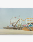 A beachside amusement park with a Ferris wheel and a roller coaster, captured beautifully for 11x14 California Canvas Print (Choose from 10+ Designs) by Offley Green. The structures are built on a pier extending over the sandy beach. The Ferris wheel is partially visible, and the roller coaster has multiple yellow tracks under a clear, cloudless sky.