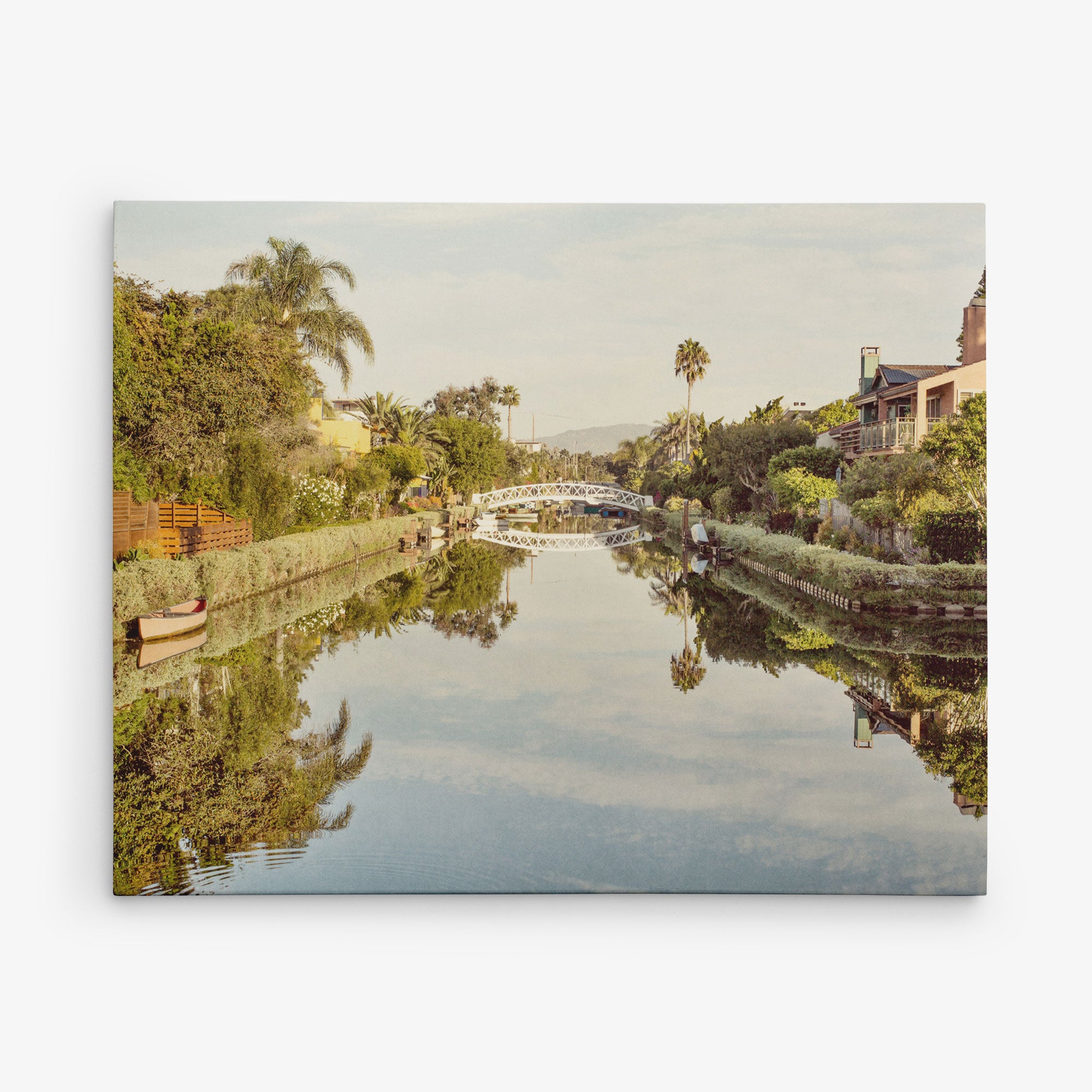 A serene canal flanked by lush greenery, houses, and palm trees is reflected in still water. A small wooden boat is docked on the left. In the distance, a decorative white bridge spans the canal under a clear sky—perfect for Offley Green&#39;s 11x14 California Canvas Print (Choose from 10+ Designs).