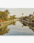 A serene canal flanked by lush greenery, houses, and palm trees is reflected in still water. A small wooden boat is docked on the left. In the distance, a decorative white bridge spans the canal under a clear sky—perfect for Offley Green's 11x14 California Canvas Print (Choose from 10+ Designs).