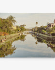 A peaceful canal lined with green trees and shrubs, with a white arched pedestrian bridge in the distance, is the perfect subject for wall art. Houses with gardens border the water, and a few small boats are visible. The clear sky and reflections in the water create a symmetrical scene ideal for Offley Green's 16x20 California Canvas Print (Choose from 10+ Designs).