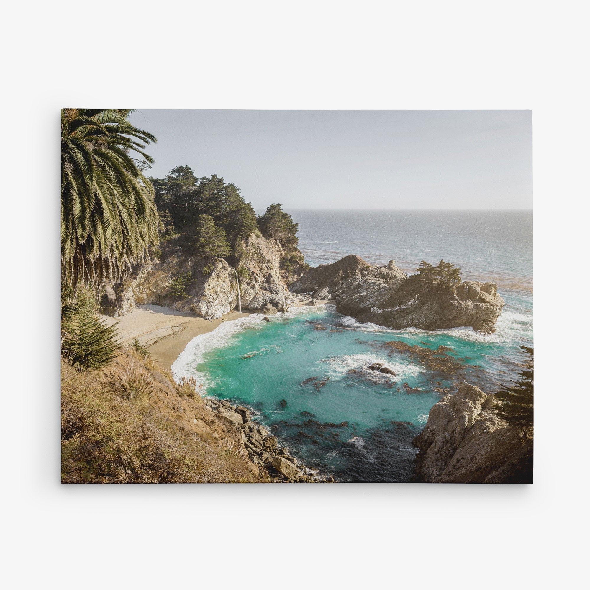 Bring the serene beauty of a secluded sandy beach surrounded by rugged rocky cliffs into your home with our 8x10 Coastal Canvas Print from Offley Green. This stunning artwork features clear turquoise waves gently lapping against the shore, lush greenery with tall palm trees adorning the cliffside, and a serene, cloudless sky meeting the horizon—perfect for wall art. Choose from over 10 beautiful designs to find the perfect match for your space.