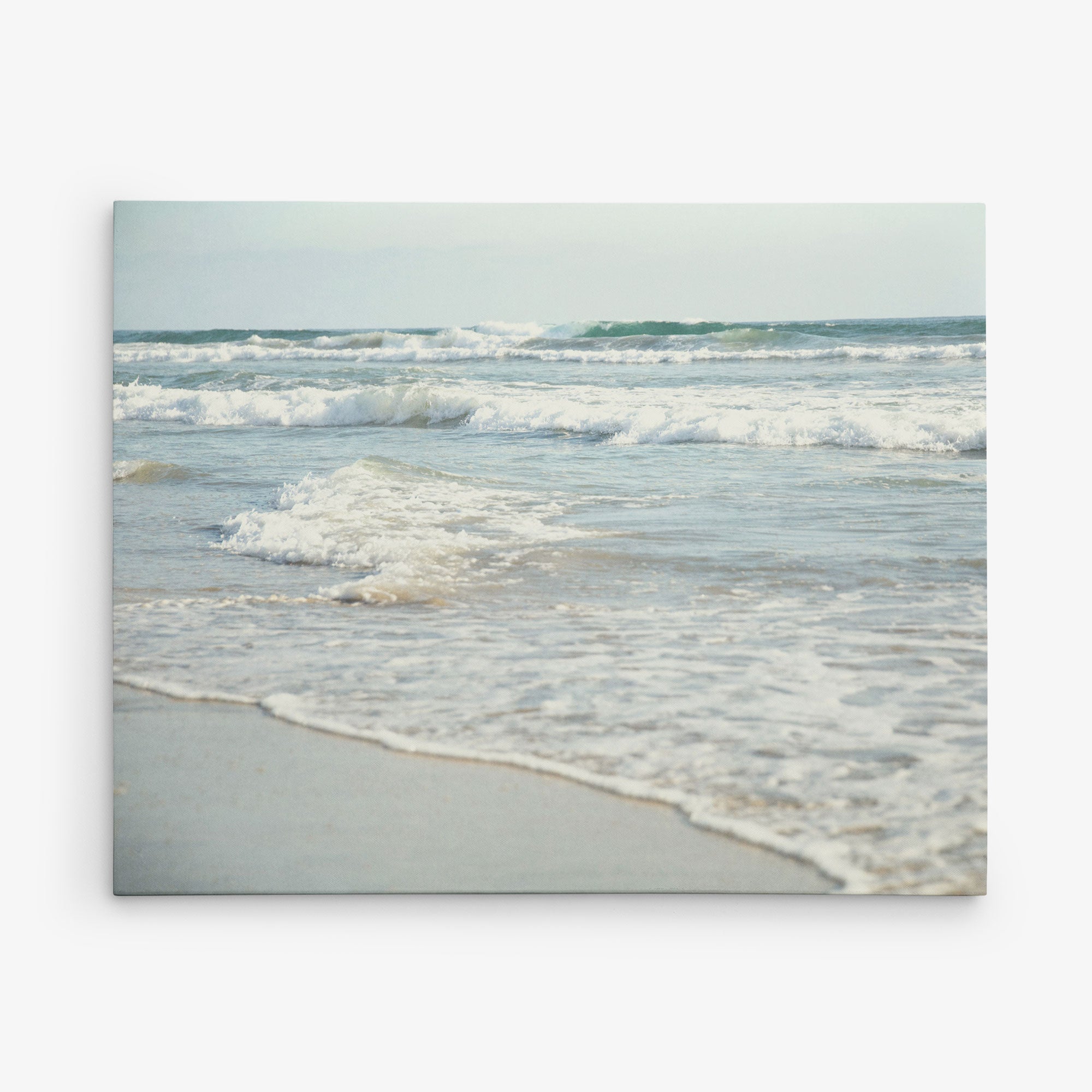 A serene beach scene showing gentle waves rolling onto a sandy shore. The water is a calm blue-green, and the horizon stretches out under a pale sky. Waves create soft white foam as they meet the sand, conveying a sense of peace and tranquility—perfect for the 8x10 Coastal Canvas Print by Offley Green (Choose from 10+ Designs).