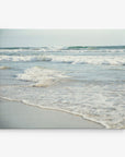 A serene beach scene showing gentle waves rolling onto a sandy shore. The water is a calm blue-green, and the horizon stretches out under a pale sky. Waves create soft white foam as they meet the sand, conveying a sense of peace and tranquility—perfect for the 8x10 Coastal Canvas Print by Offley Green (Choose from 10+ Designs).