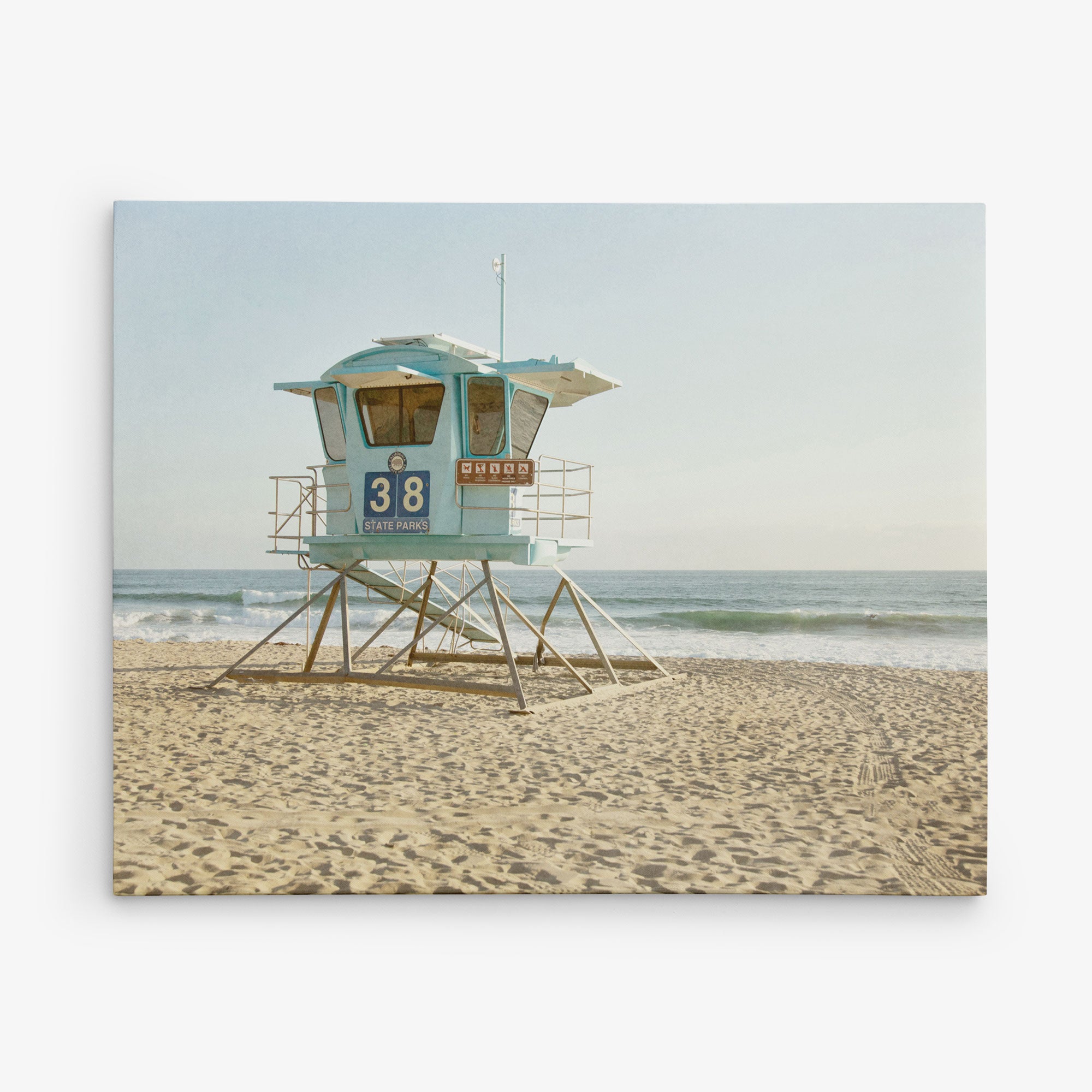 A solitary blue lifeguard tower labeled &quot;38&quot; stands on a sandy beach with the ocean in the background. The sky is clear, and the waves gently approach the shore. This calm, sunny day at the beach is perfect for Offley Green&#39;s 11x14 California Canvas Print (Choose from 10+ Designs).