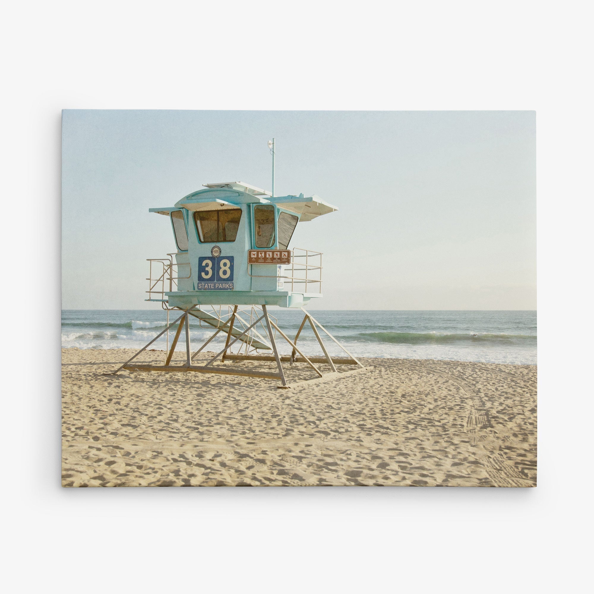 A serene 30x40 California Canvas Print by Offley Green features a light blue lifeguard tower numbered &quot;38&quot; on a sandy beach, with the ocean and gentle waves in the background. The clear sky hints at a calm, sunny day, making it an ideal piece of wall art reminiscent of classic California prints. Choose from over 10 stunning designs to capture this perfect beach scene in your home.