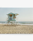 A solitary blue lifeguard tower labeled "38" stands on a sandy beach with the ocean in the background. The sky is clear, and the waves gently approach the shore. This calm, sunny day at the beach is perfect for Offley Green's 11x14 California Canvas Print (Choose from 10+ Designs).