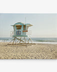 A blue lifeguard tower labeled "38" stands on a sandy beach with the ocean waves gently rolling in the background. The sky is clear and the sun is shining, casting a soft, warm light on this perfect California scene, ideal for an Offley Green 16x20 California Canvas Print (Choose from 10+ Designs).