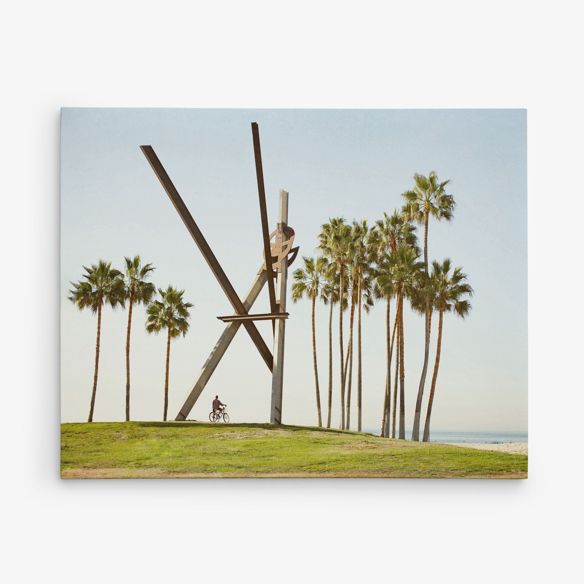 A large, abstract metal sculpture sits on a grassy hill near a beach, surrounded by tall palm trees. A person is riding a bicycle in front of the sculpture. The sky is clear and the ocean is visible in the background, creating a scene perfect for Offley Green&#39;s 11x14 California Canvas Print (Choose from 10+ Designs).